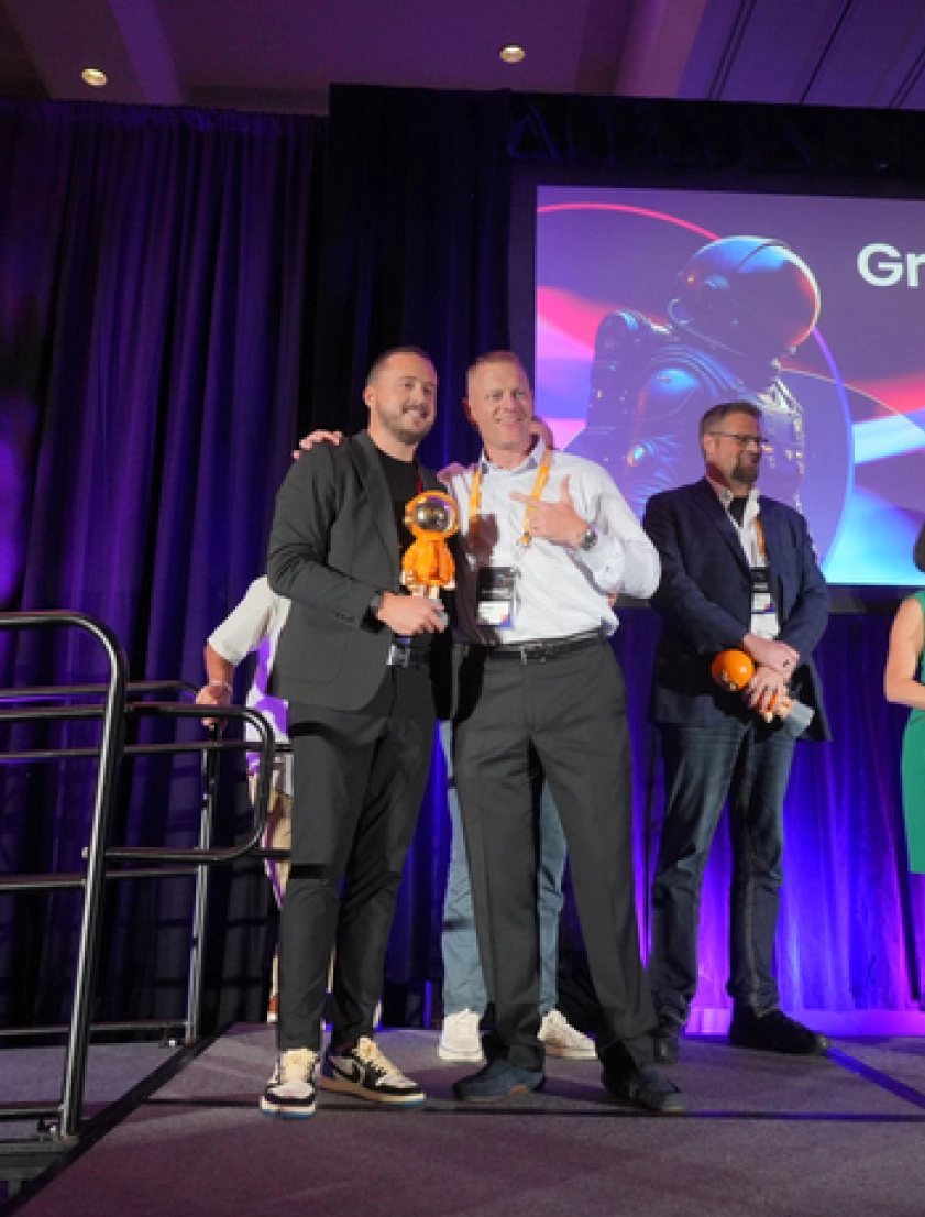 Two men on stage smiling, one holding an orange astronaut figurine; a third man and a woman stand in the background near a screen displaying an astronaut image.