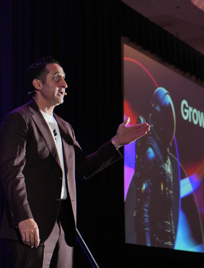 Man in a dark suit speaking and gesturing with his right hand in front of a screen showing a futuristic astronaut figure and the word 'Grow'.