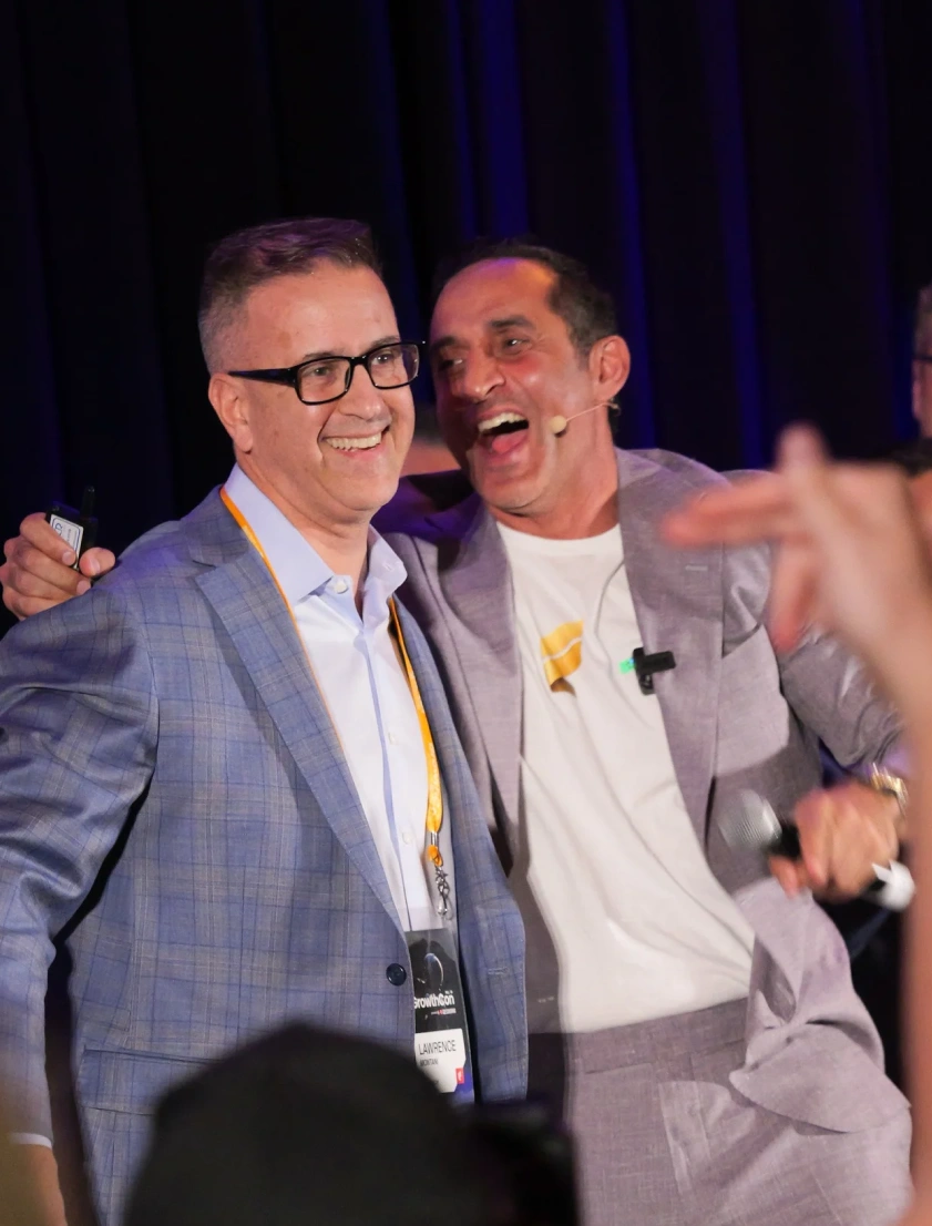 Two men smiling and embracing at an event, one wearing glasses and a blue checkered suit, the other in a light gray suit with a microphone headset.