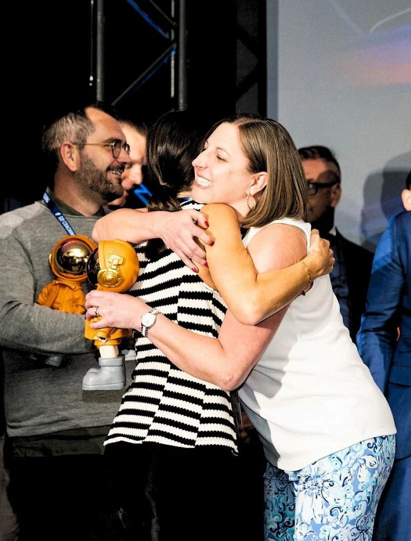 Two women embracing warmly while a man nearby holds a small golden astronaut figurine during an indoor event.