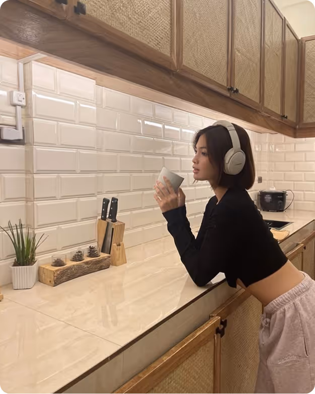 Young woman wearing headphones, leaning on a kitchen counter holding a cup.