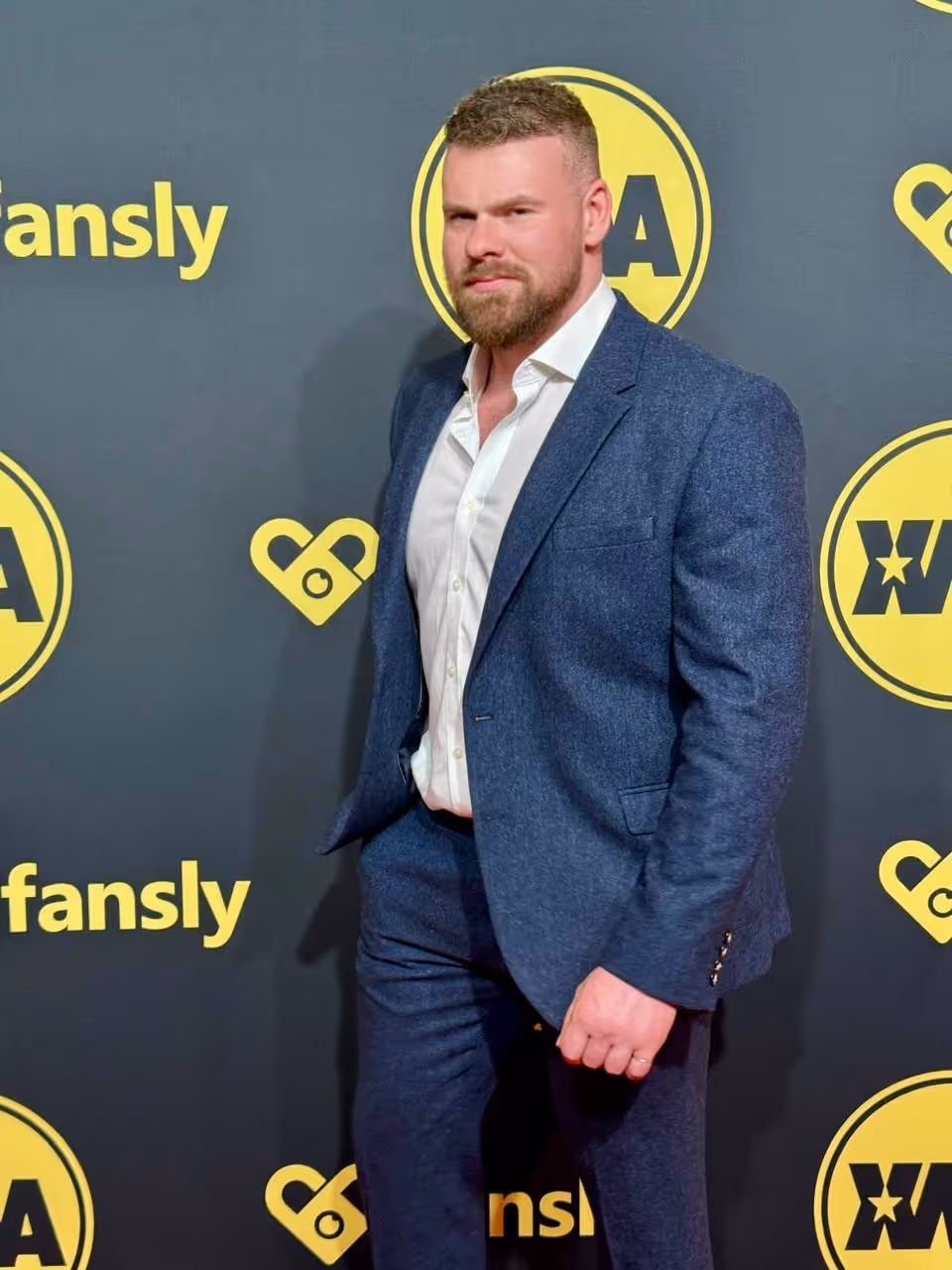 Man with beard wearing a blue suit and white shirt posing in front of a Fansly event backdrop.