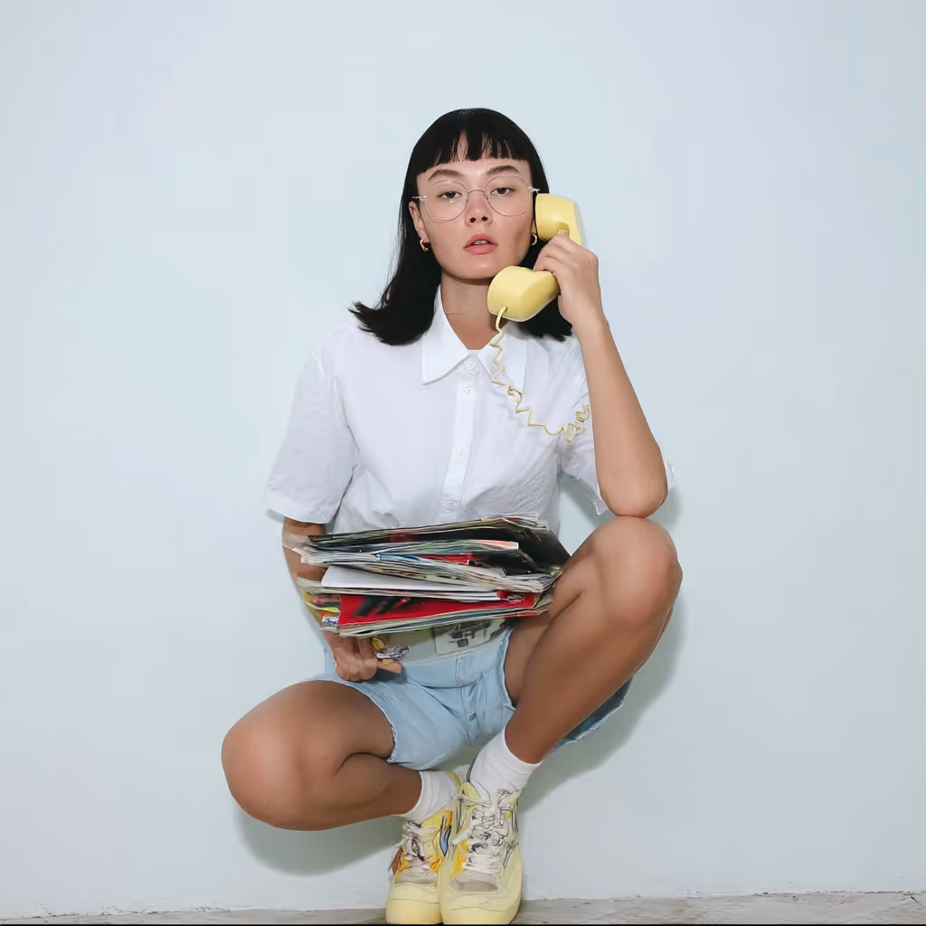 Woman with glasses in white shirt and shorts squatting while holding a yellow telephone handset and a stack of magazines.