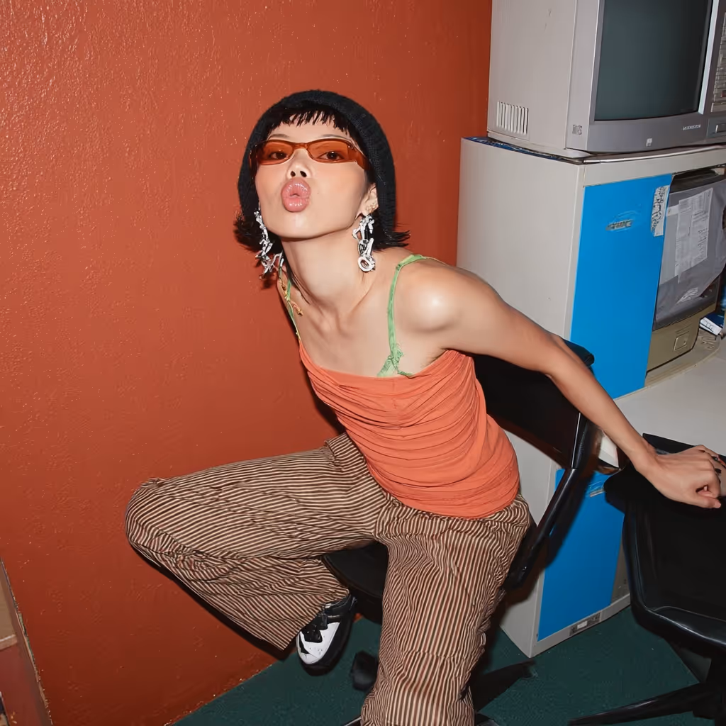 Person with short black hair wearing orange sunglasses, a black beanie, orange top, and striped pants sitting on a chair against an orange wall.