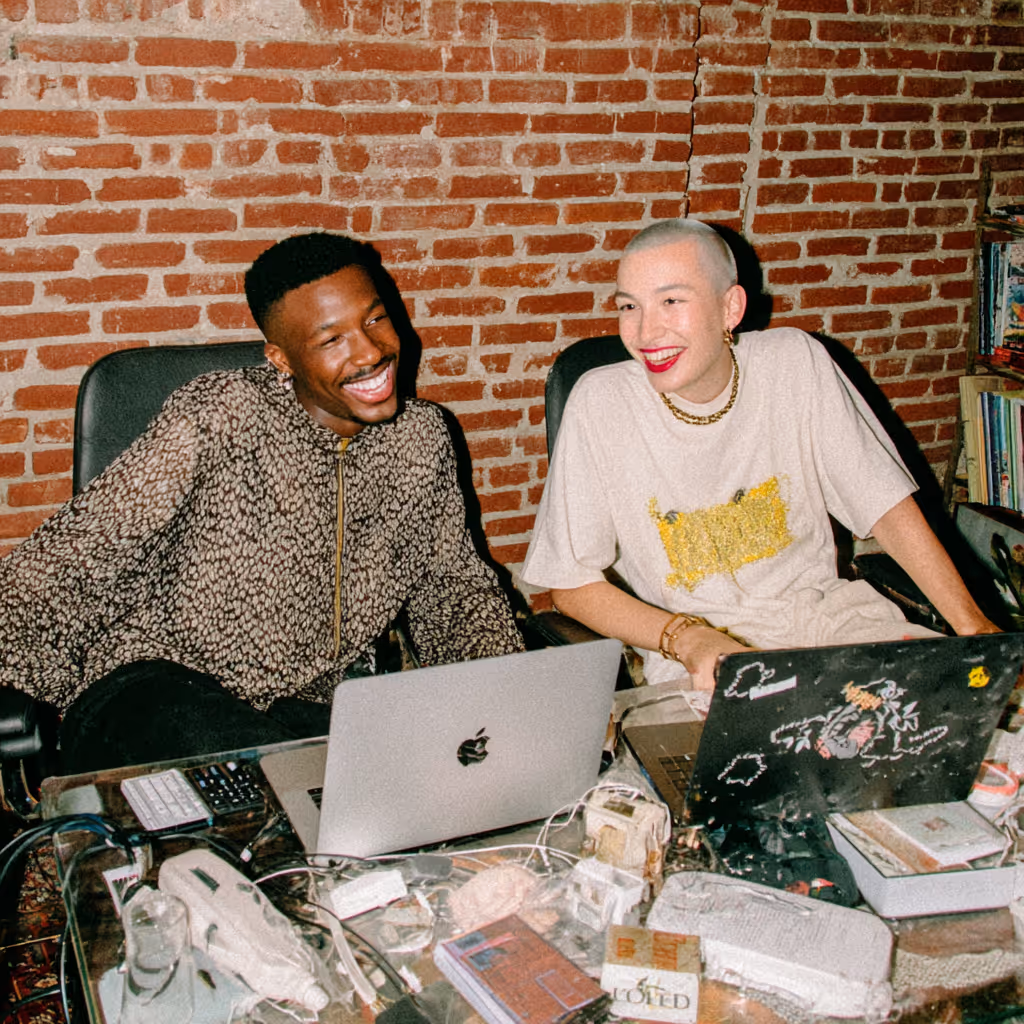 Two people sitting at a cluttered glass table with laptops, smiling and engaged in conversation against a brick wall background.