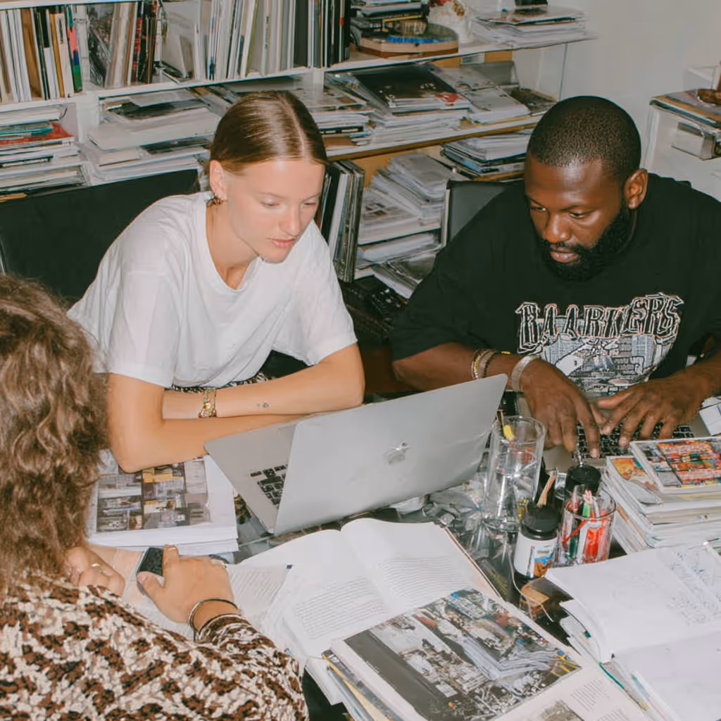 Three people working together at a cluttered desk with open books, papers, and a laptop in a room filled with shelves of books and magazines.
