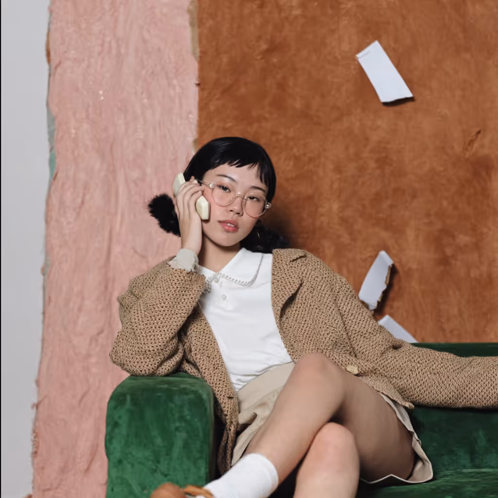 Young woman with glasses, wearing a beige knitted cardigan and skirt, sitting on a green sofa and talking on a retro phone.