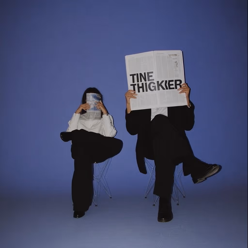 Two people sitting on transparent chairs against a blue background, each with one leg crossed and hiding their faces behind newspapers.