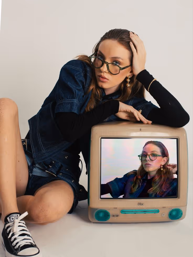 Woman with glasses leaning on a vintage iMac displaying her own image wearing the same outfit and glasses.