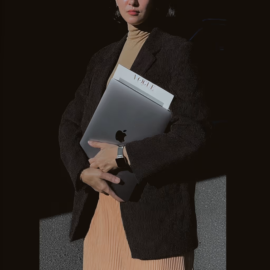 Person in dark blazer holding a closed MacBook laptop with a Vogue magazine tucked inside.