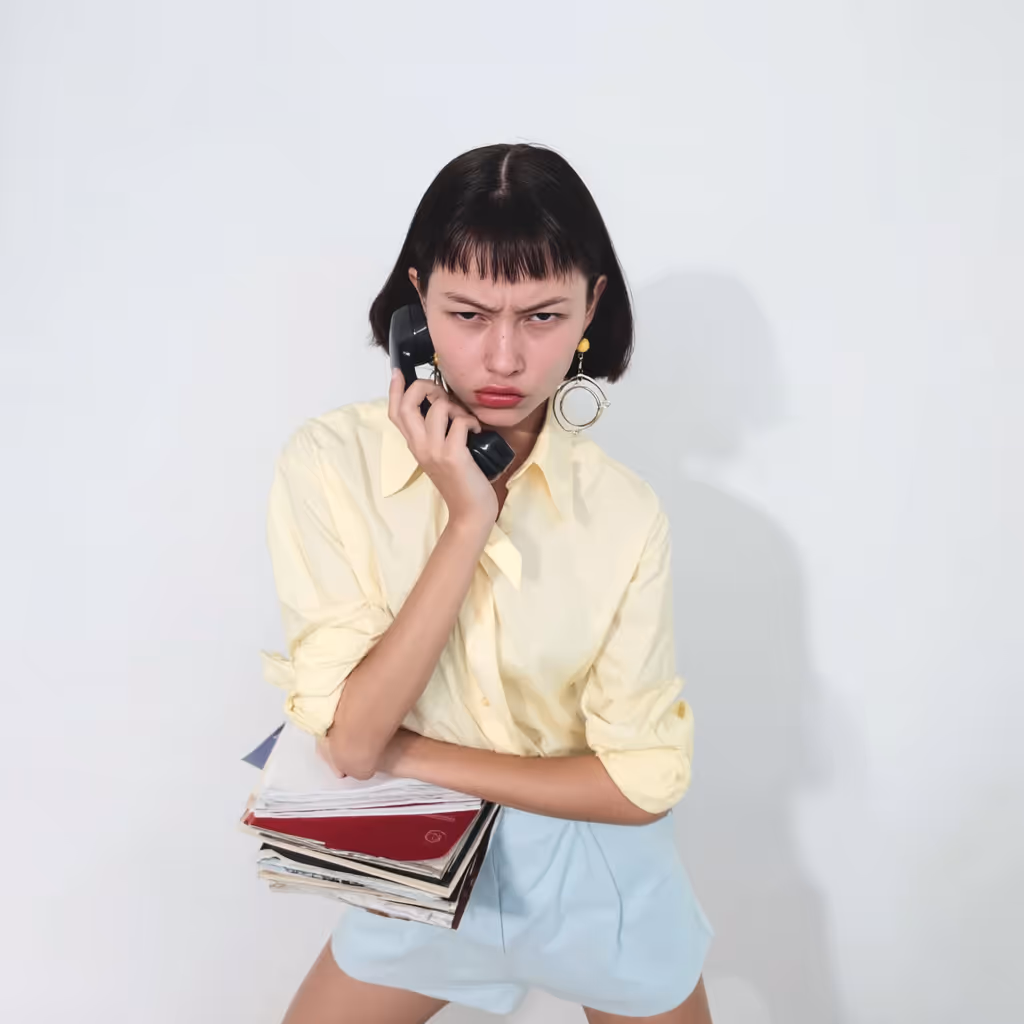 Woman with short dark hair, wearing a yellow shirt and light blue skirt, holding a stack of documents and a phone receiver with a frustrated expression.