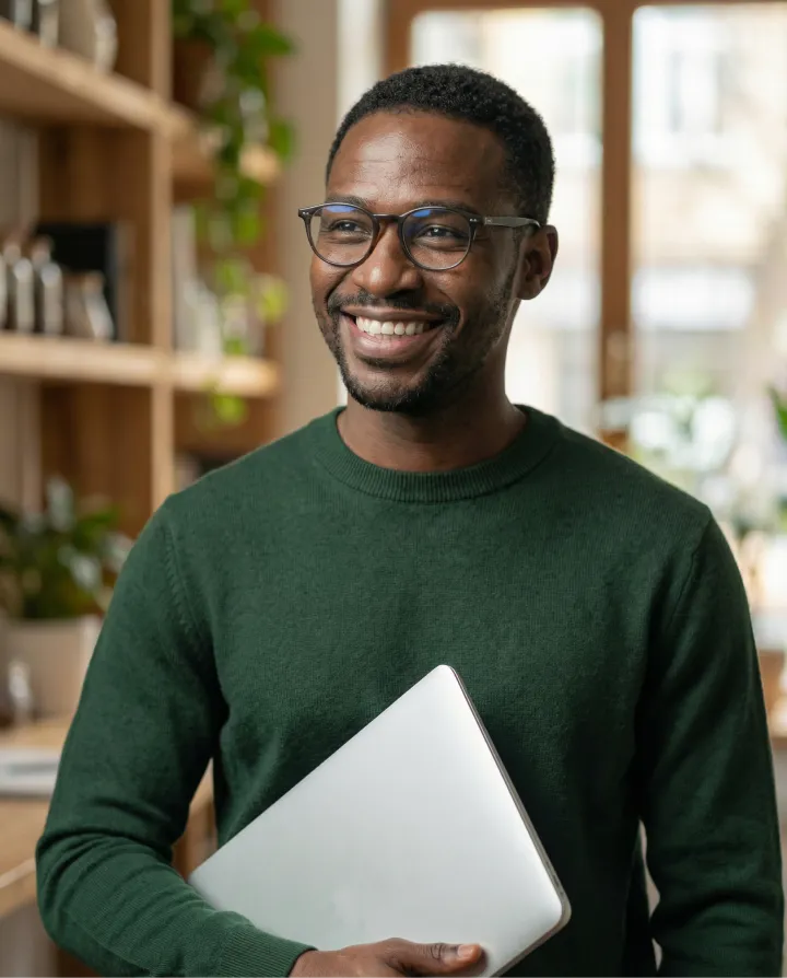 Smiling man wearing glasses and a green sweater holding a closed laptop in a cozy room with plants.