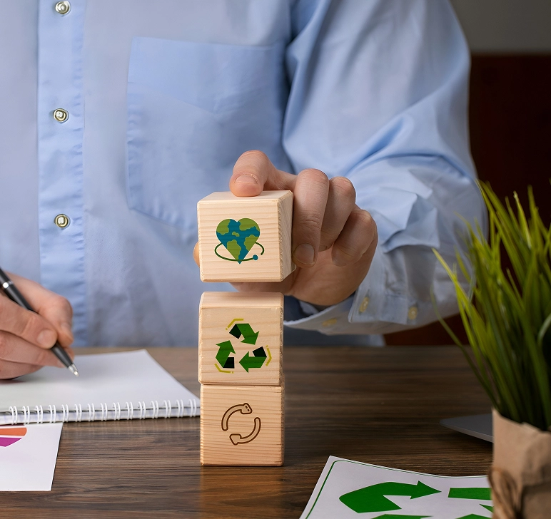 Person stacking wooden blocks with eco-friendly symbols including recycling arrows and Earth-shaped heart.