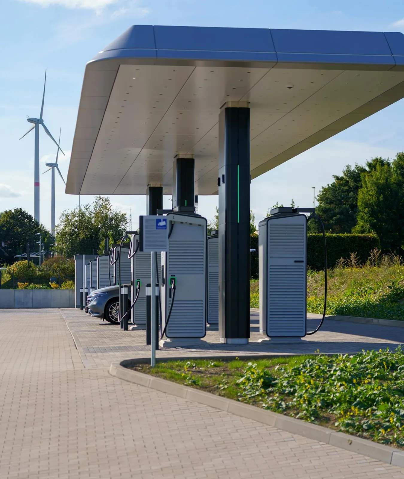 Modern electric vehicle charging station with multiple chargers under a blue canopy and wind turbines in the background.