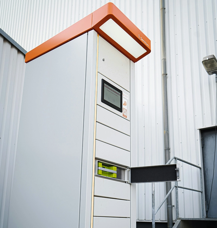 Tall white battery storage unit with an orange top and a small screen, one drawer partially open revealing green batteries.