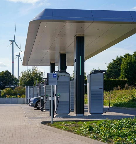 Electric vehicle charging station with multiple charging units and parked cars under a canopy, wind turbines and greenery in the background.