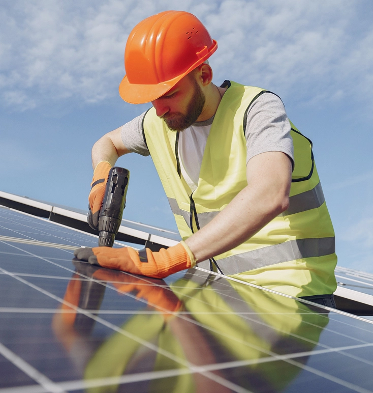 Construction worker wearing an orange hard hat and yellow safety vest installing solar panels with a drill under a partly cloudy sky.