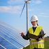 Technician in hard hat and safety vest inspecting solar panels with a laptop near a wind turbine.