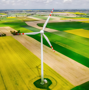 Aerial view of a single wind turbine standing in the middle of green and yellow agricultural fields under a partly cloudy sky.