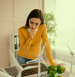Woman in yellow sweater examining a solar panel and wind turbine models on a table with small plants.