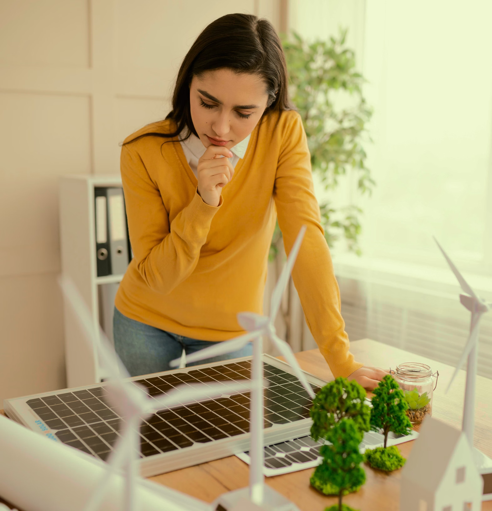 Woman in yellow sweater examining wind turbine models and solar panels on a table with miniature trees.