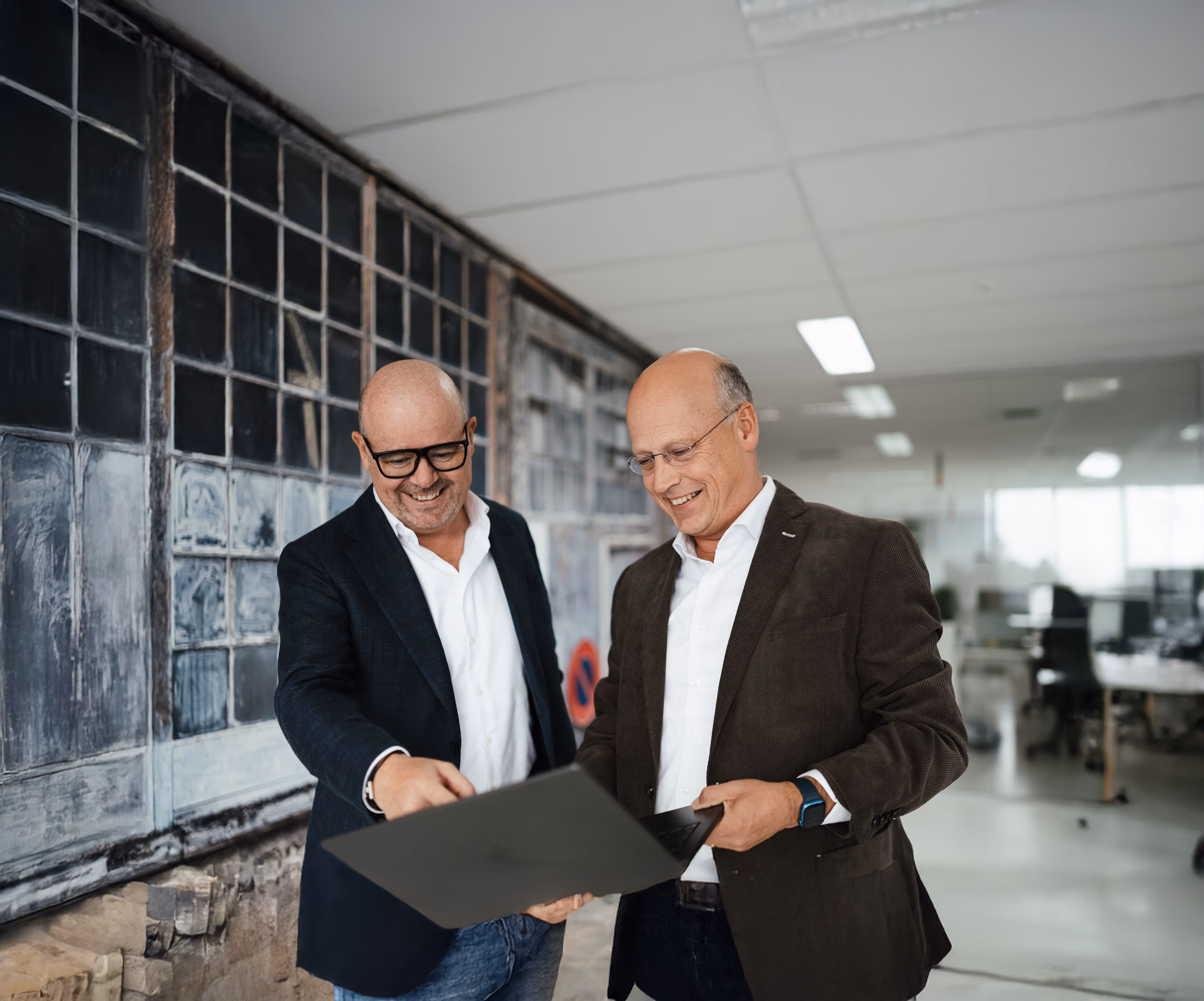 Two bald men in business attire smiling and looking at a black folder or laptop in a modern office with large windows behind them.