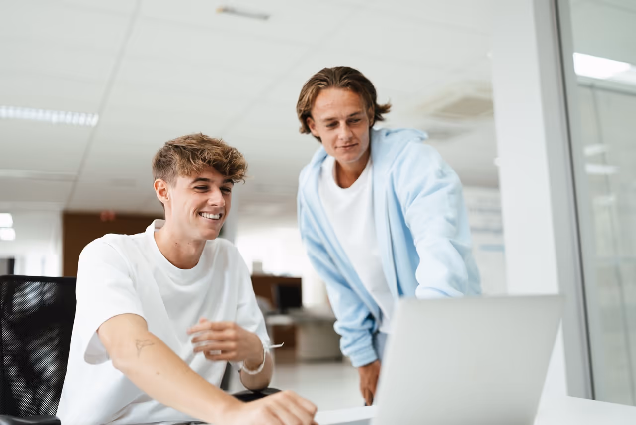 Two young men smiling and looking at a laptop in a bright office setting.