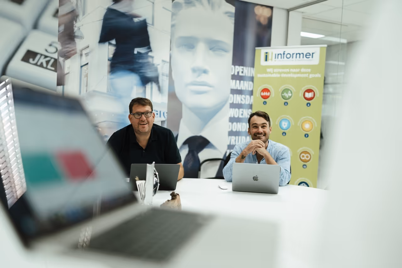 Two men sitting at a white conference table with laptops, smiling in a modern office with glass walls and a large statue face poster in the background.