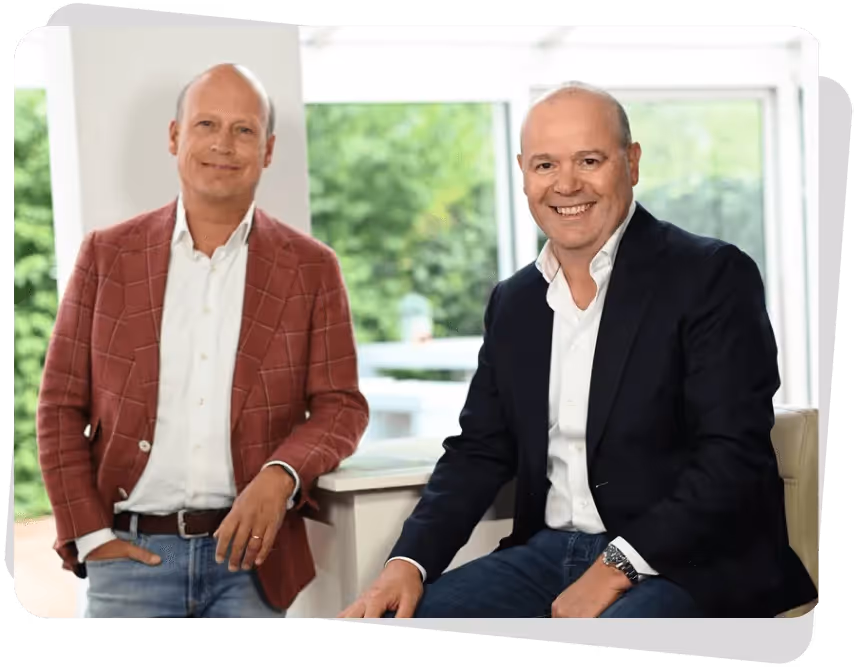 Two smiling men seated indoors by a window with a green outdoor background; one wearing a red checkered blazer and the other in a black blazer.