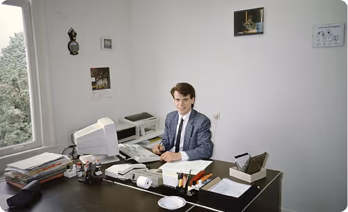 Man in a suit sitting at a cluttered office desk with computer and papers, looking at the camera.