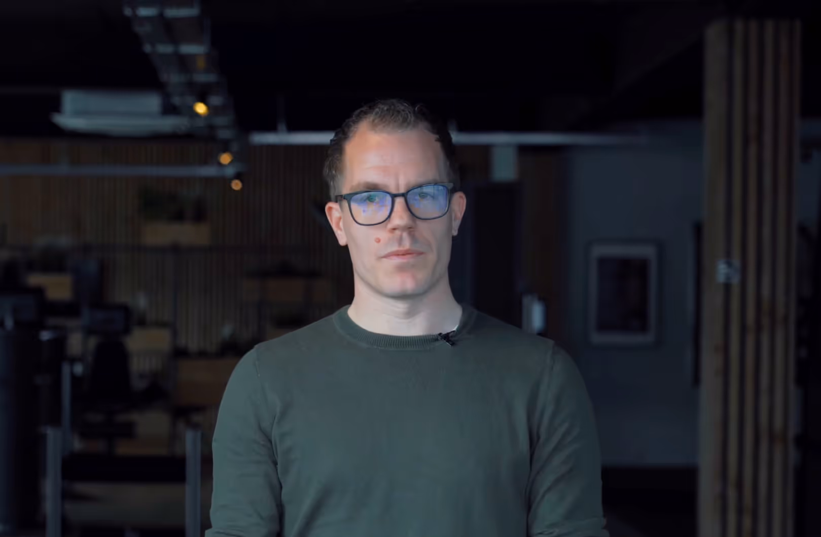 Man with glasses wearing a green sweater standing in a dimly lit modern office space.