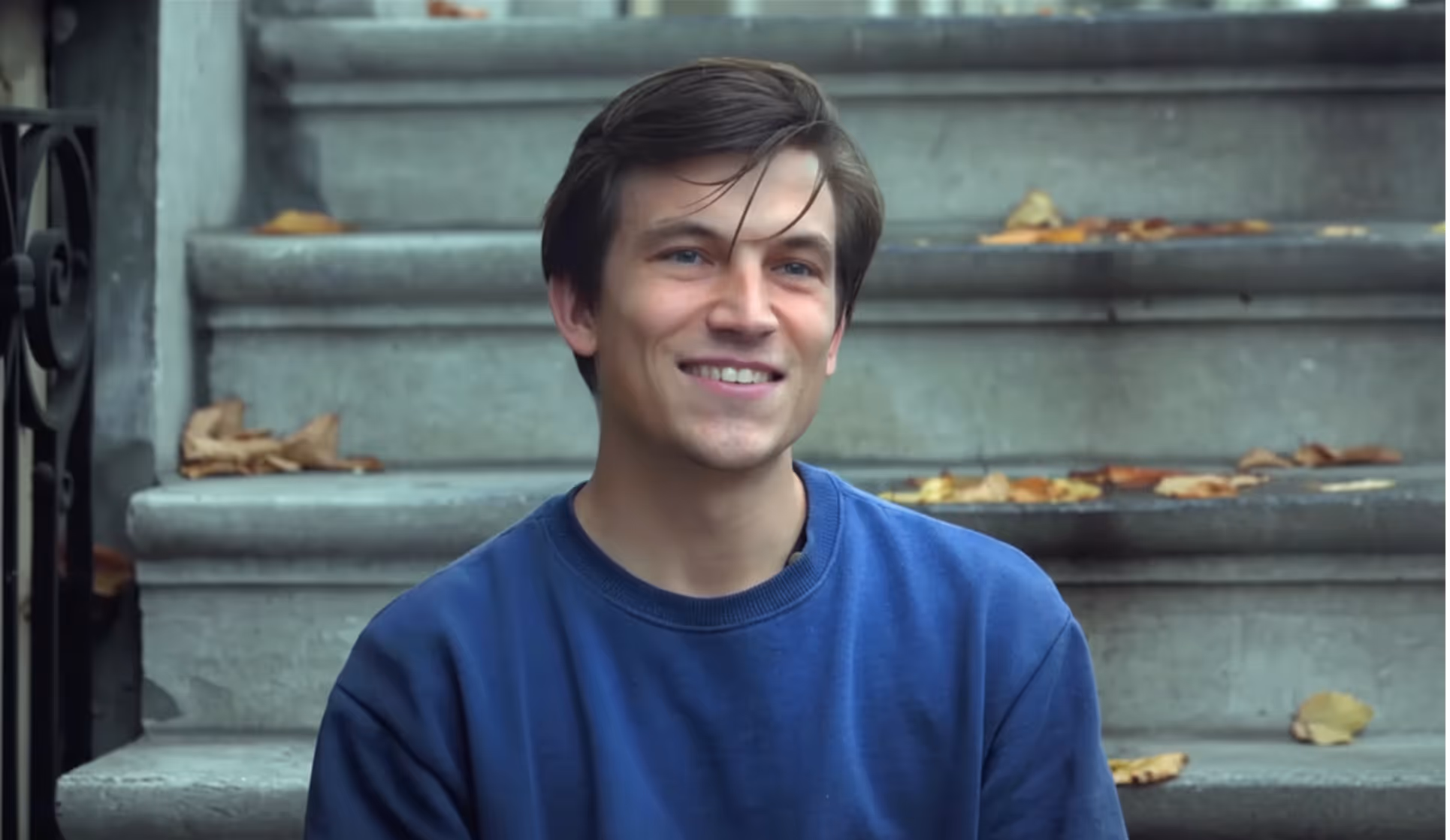 Smiling young man wearing a blue sweater sitting on outdoor stone steps with autumn leaves scattered around.