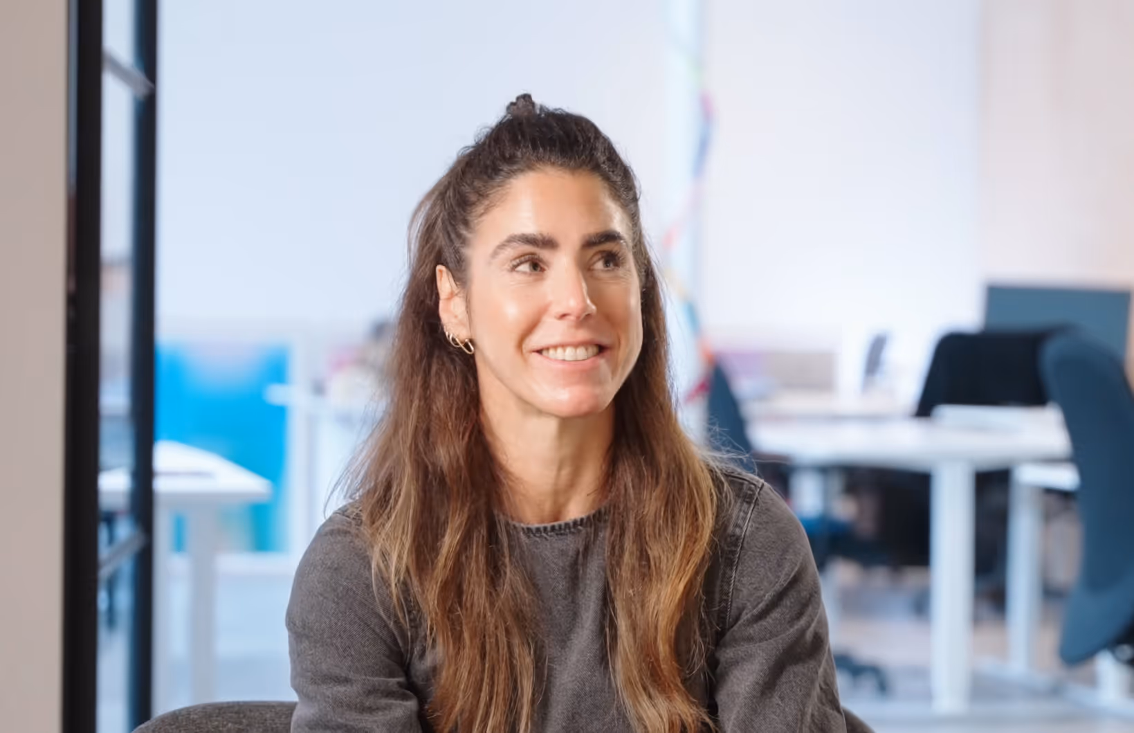 Smiling woman with long hair in a gray sweater sitting in a modern office.