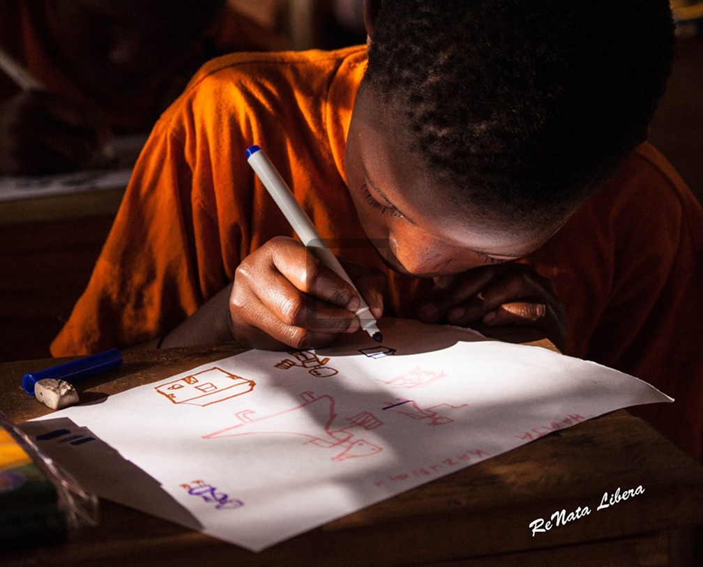 Child in orange shirt drawing on paper with a black marker in a dimly lit room.