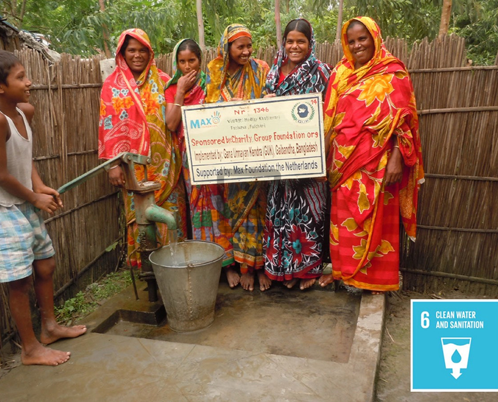 Group of women in colorful saris and a boy pumping water from a hand pump into a bucket, with a sign showing support from charity organizations in Bangladesh and the Netherlands.