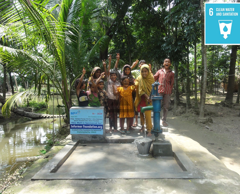 A group of children gathered around a blue hand pump well with water flowing into a bucket, surrounded by green trees and plants.