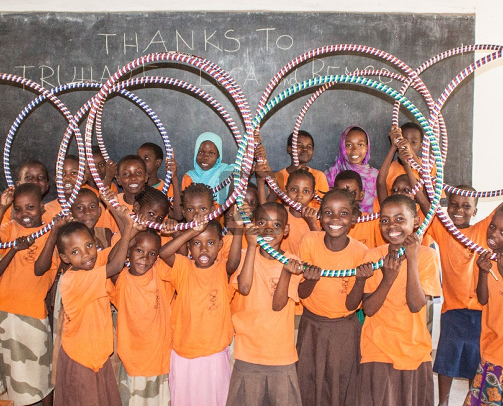 Group of smiling children in orange shirts holding colorful hula hoops in front of a chalkboard.