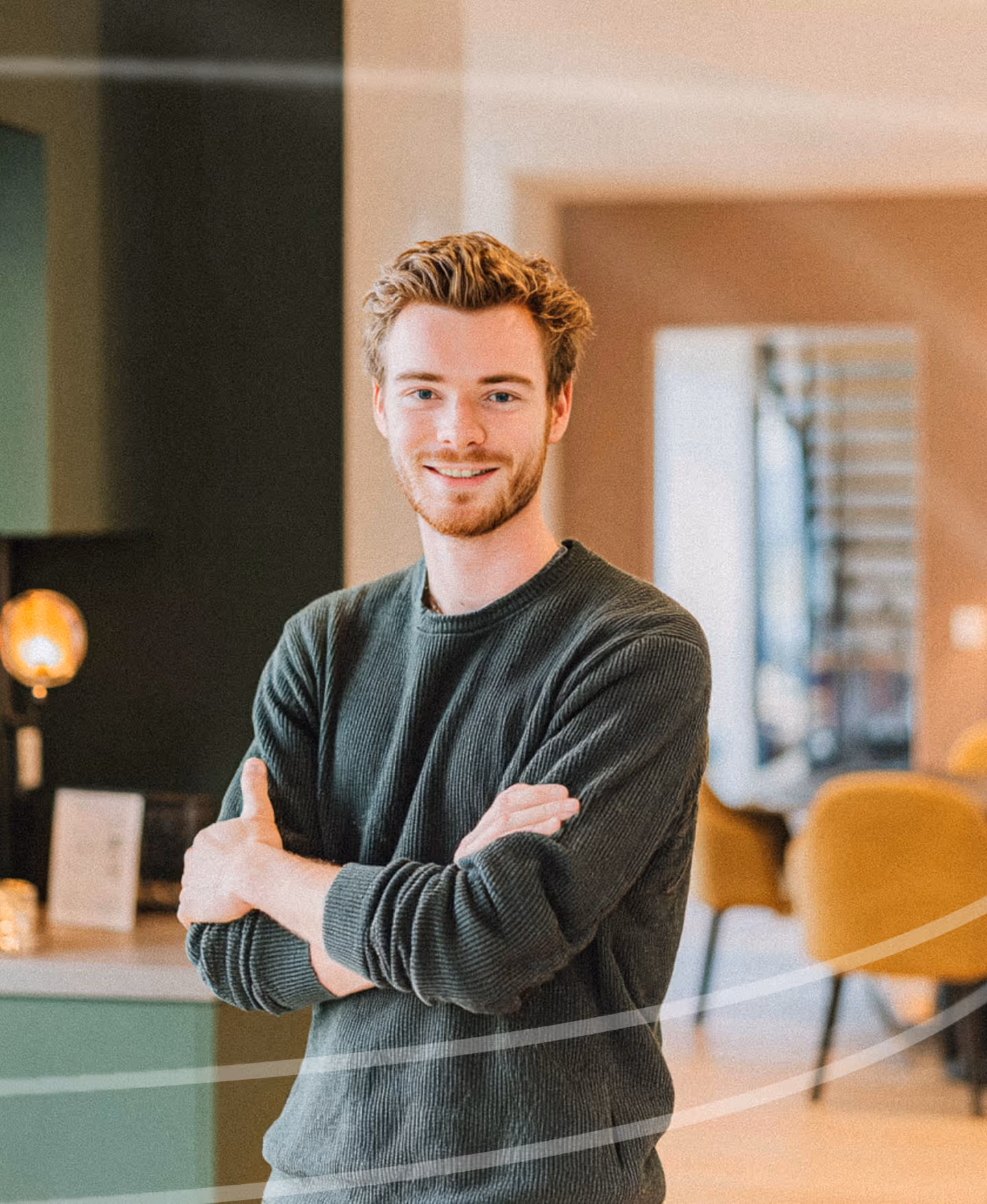 Smiling young man with crossed arms standing indoors in a modern room with chairs and a lamp in the background.