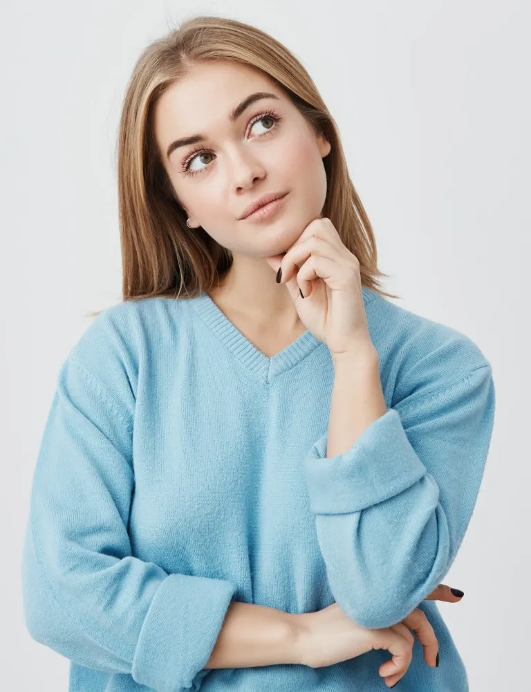 Young woman in a light blue sweater looking thoughtfully upwards with her hand resting near her chin.