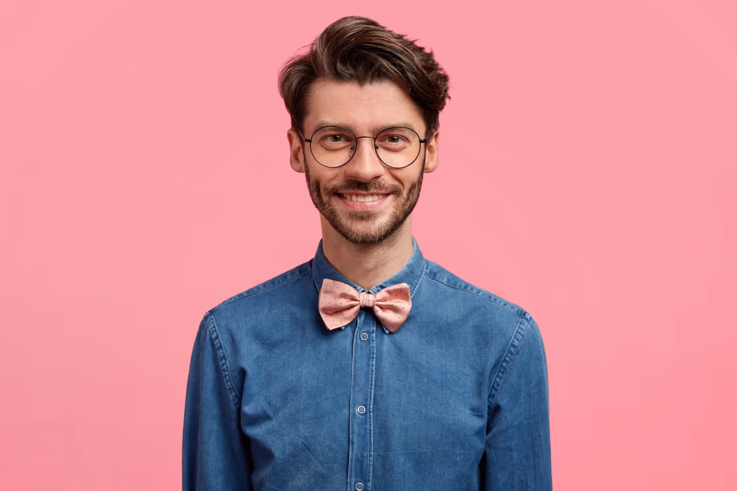 Smiling man with brown hair and glasses wearing a blue button-up shirt and pink bow tie against a pink background.