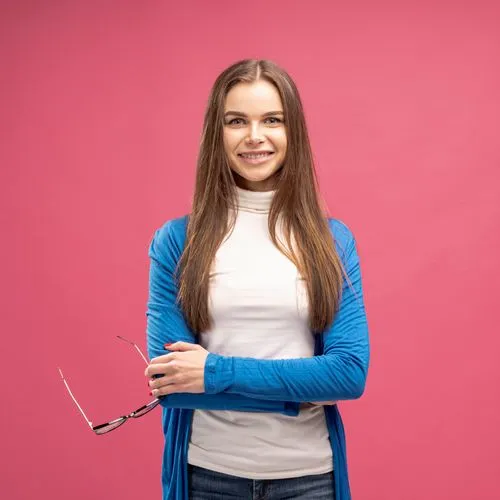 Smiling woman with long brown hair wearing a blue cardigan and white turtleneck, holding glasses, against a pink background.