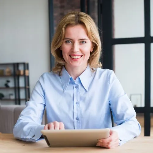 Smiling woman in a light blue shirt using a tablet at a wooden table indoors.