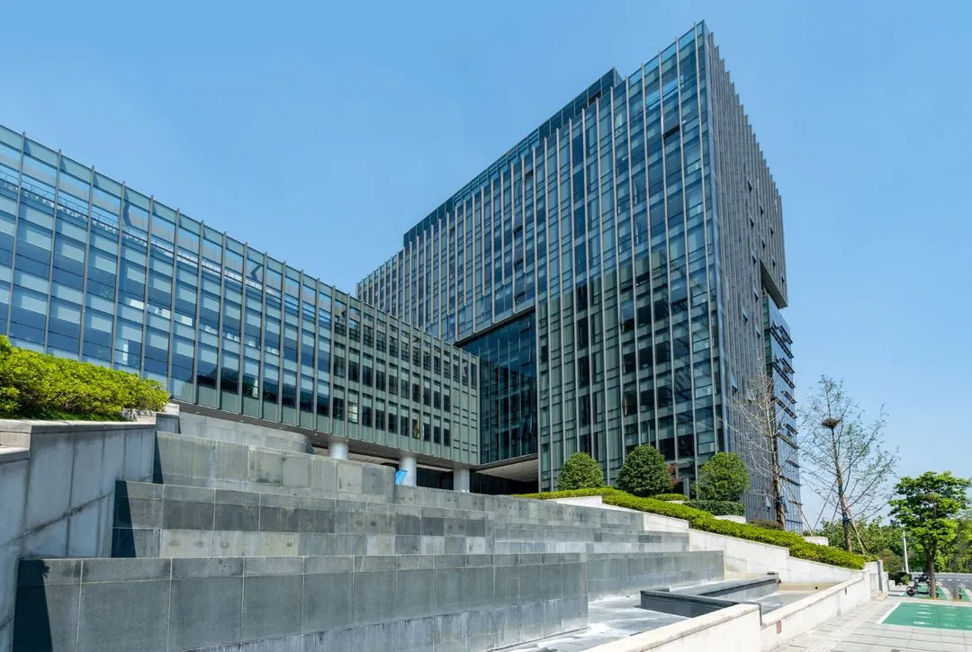 Modern glass office building with concrete steps and greenery under a clear blue sky.