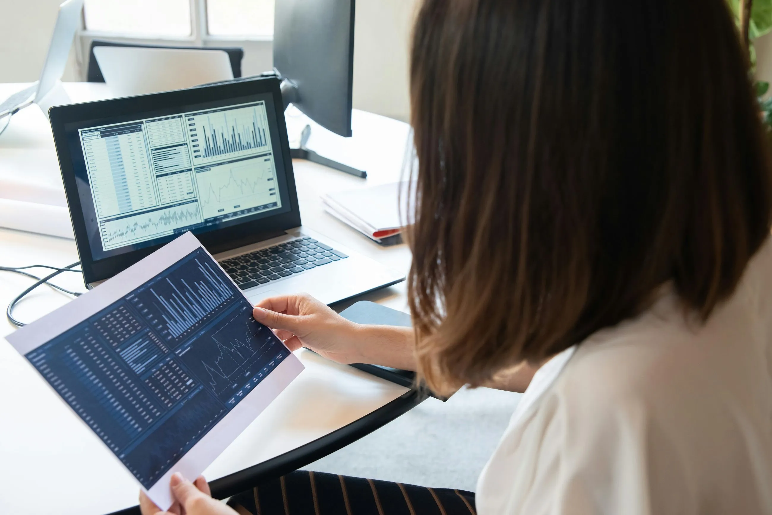 Person analyzing printed and digital financial charts with graphs and data at a desk.