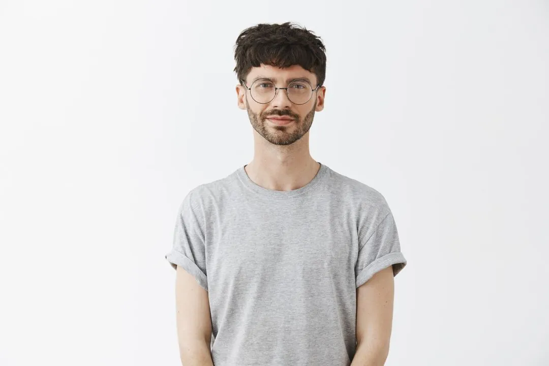 Young man with short dark hair, beard, and round glasses wearing a light grey t-shirt standing against a white background.