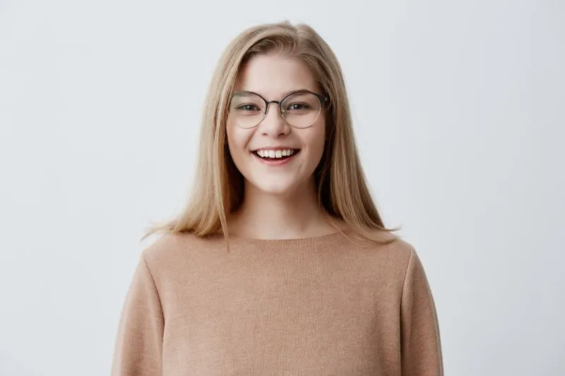 Smiling young woman with blonde hair and glasses wearing a light brown sweater against a plain gray background.