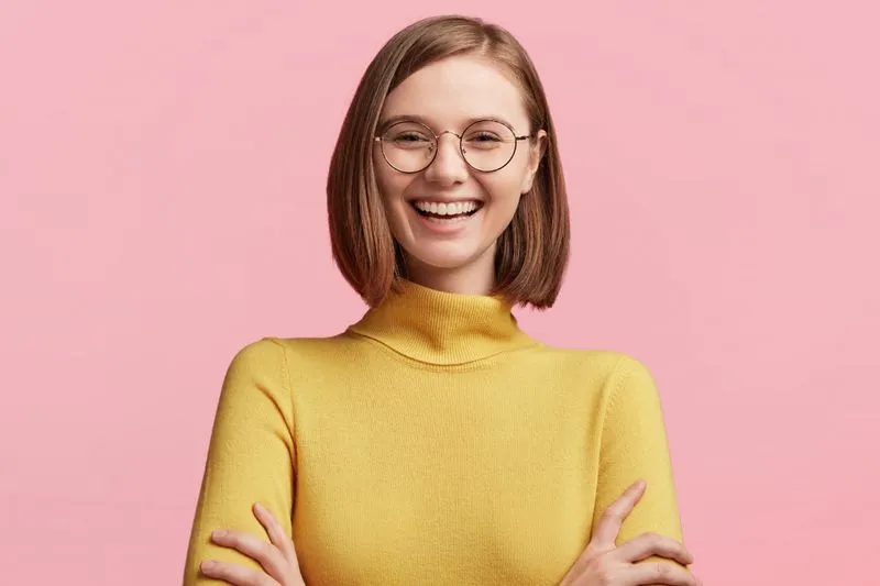 Smiling young woman with short brown hair and glasses wearing a yellow turtleneck sweater against a pink background.