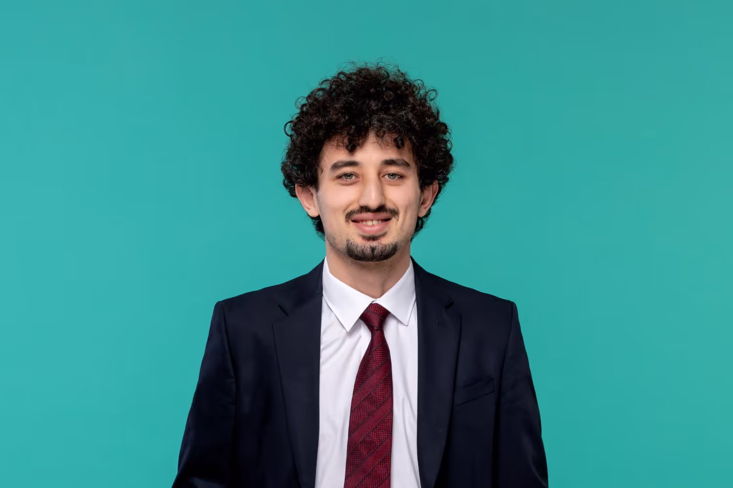 Smiling man with curly hair wearing a dark suit, white shirt, and red tie against a teal background.