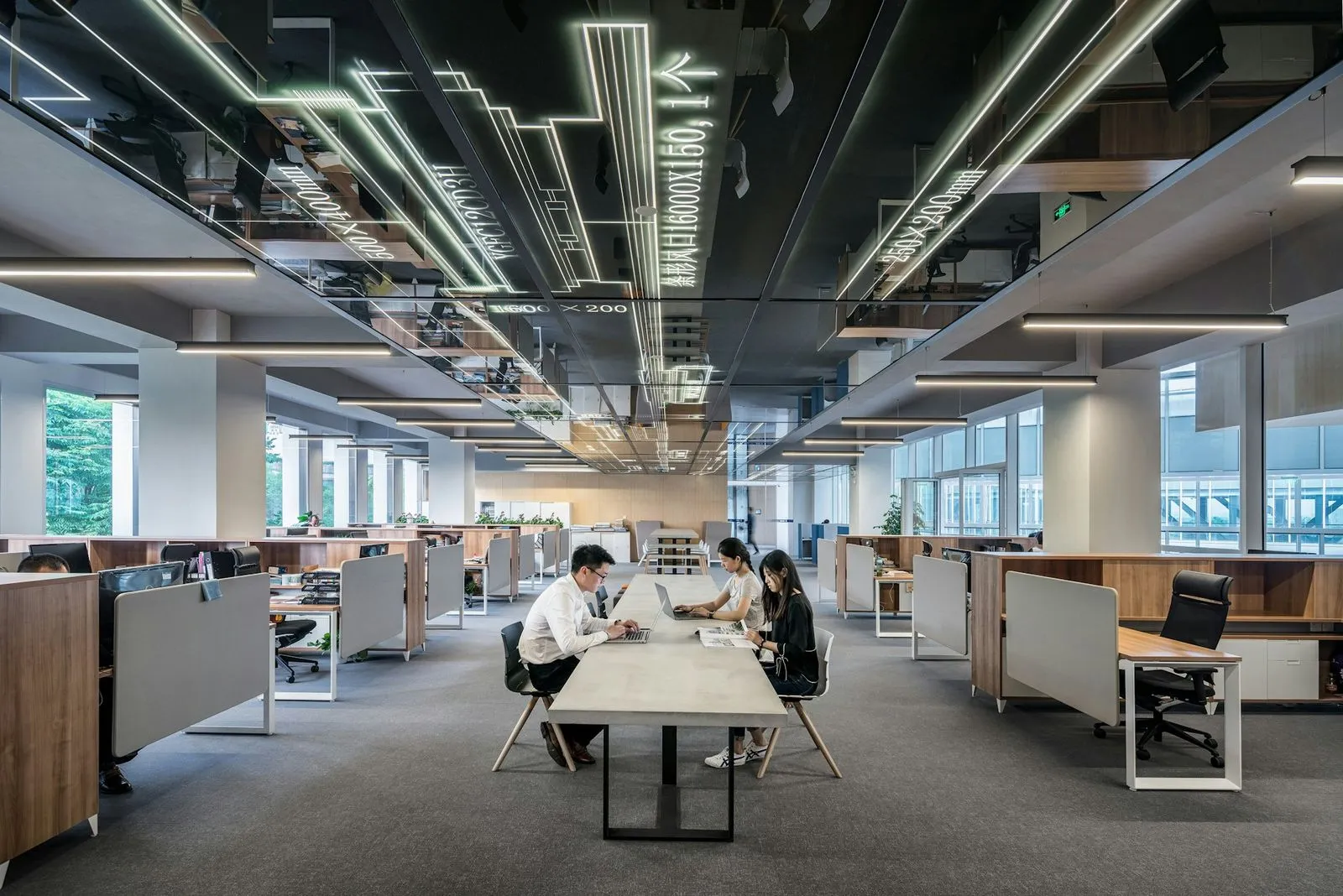 Modern open office space with rows of workstations and three people working at a long central table with laptops and documents.