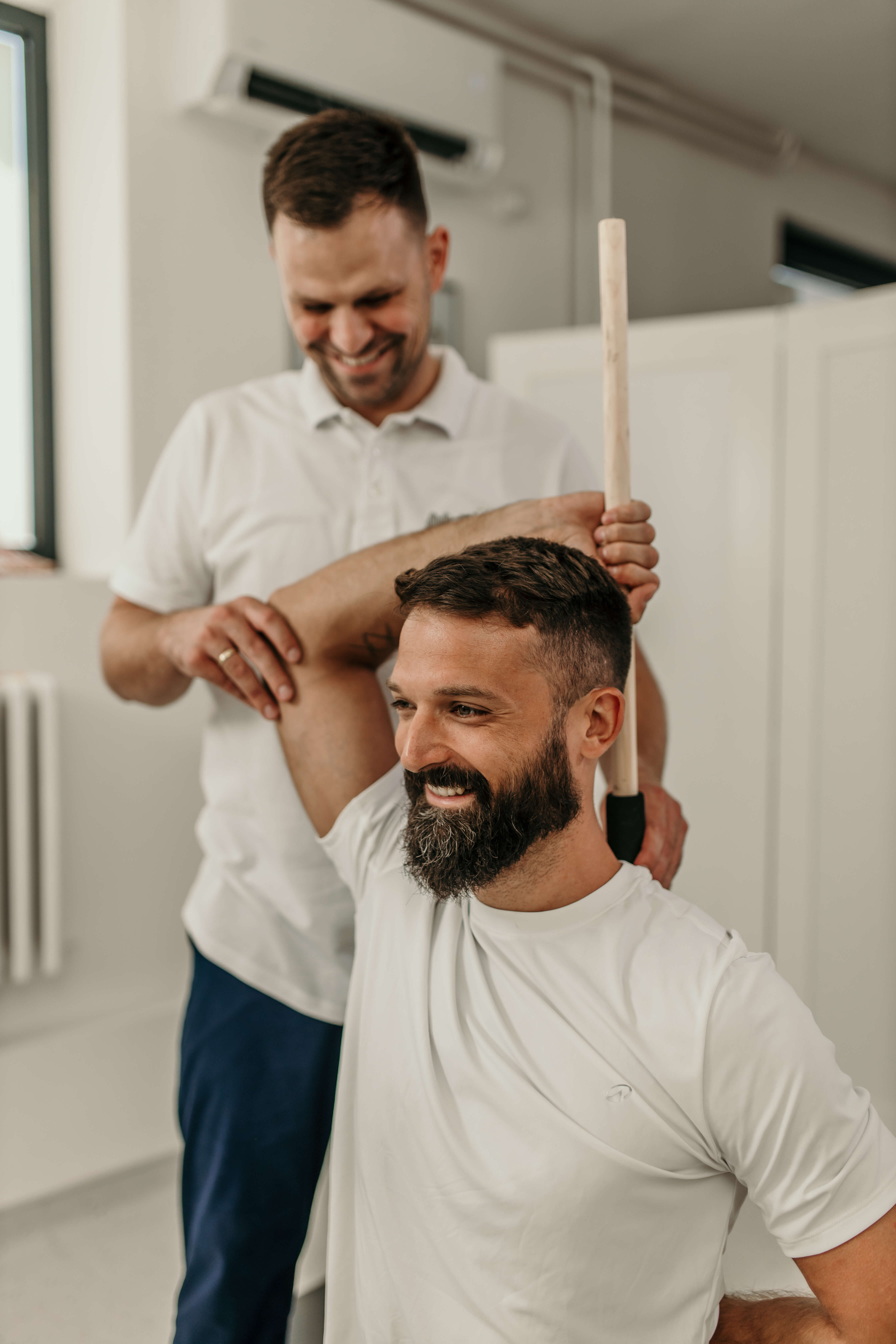 Physiotherapist helping a smiling man stretch his arm using a wooden stick in a bright room. Treasure Coast Physical Therapy and Fitness.